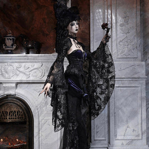 Model in a gothic black lace robe with purple corset, feathered hat, holding a small dark rose by a fireplace.