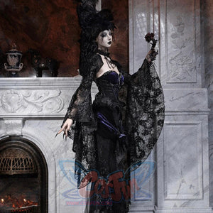 Model in a gothic black lace robe with purple corset, feathered hat, holding a small dark rose by a fireplace.