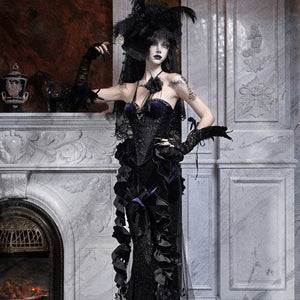 Model in a gothic black ruffled mermaid skirt with feathered hat, standing beside a marble fireplace.