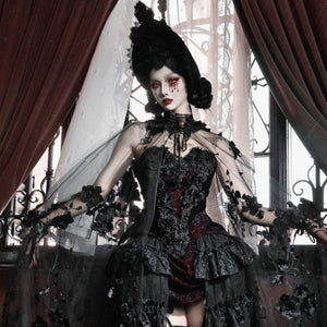 Model wearing gothic black velvet corset bustier top with red brocade, sheer veil, black floral arm accents, and dark crown.