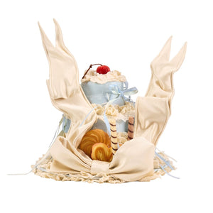 Cookie-shaped cake hat flat lay with satin wing details, lace trim, and dessert accents