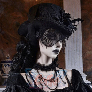 A model wearing a black Gothic Victorian flat-top hat with embroidered rose detail, black feathers, and a black lace veil, styled with a velvet gothic dress and layered necklaces