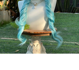 Close-up view of peacock teal blue lolita wig showing long braided pigtails on a mannequin outdoors, displayed on grass with potted plants and greenery in the background