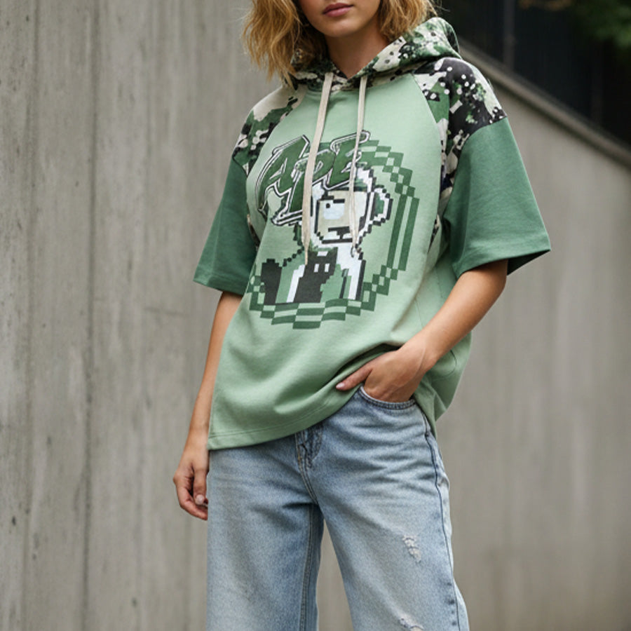Person wearing a green graphic hoodie and jeans against a concrete wall.