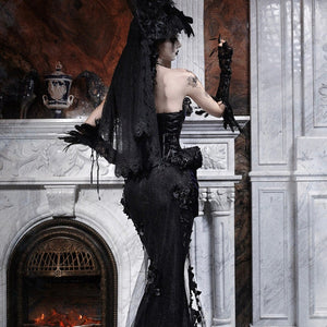 Back view of a model in a gothic black mermaid skirt with a long lace veil, standing near a marble fireplace.