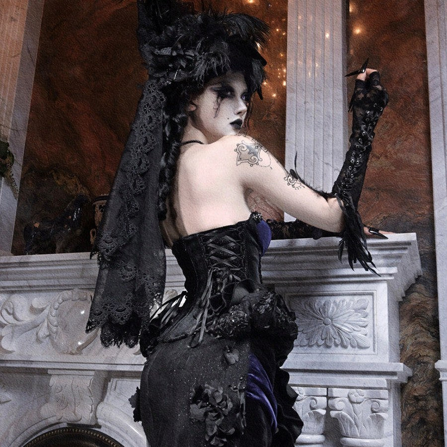 Back view of a model in a gothic black corset with lace overlay skirt, wearing a feathered hair piece.