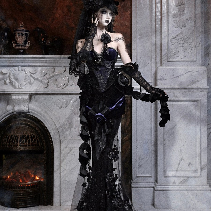 Model wearing a gothic black lace mermaid skirt and feathered hat, shown in a three-quarter pose by a fireplace.