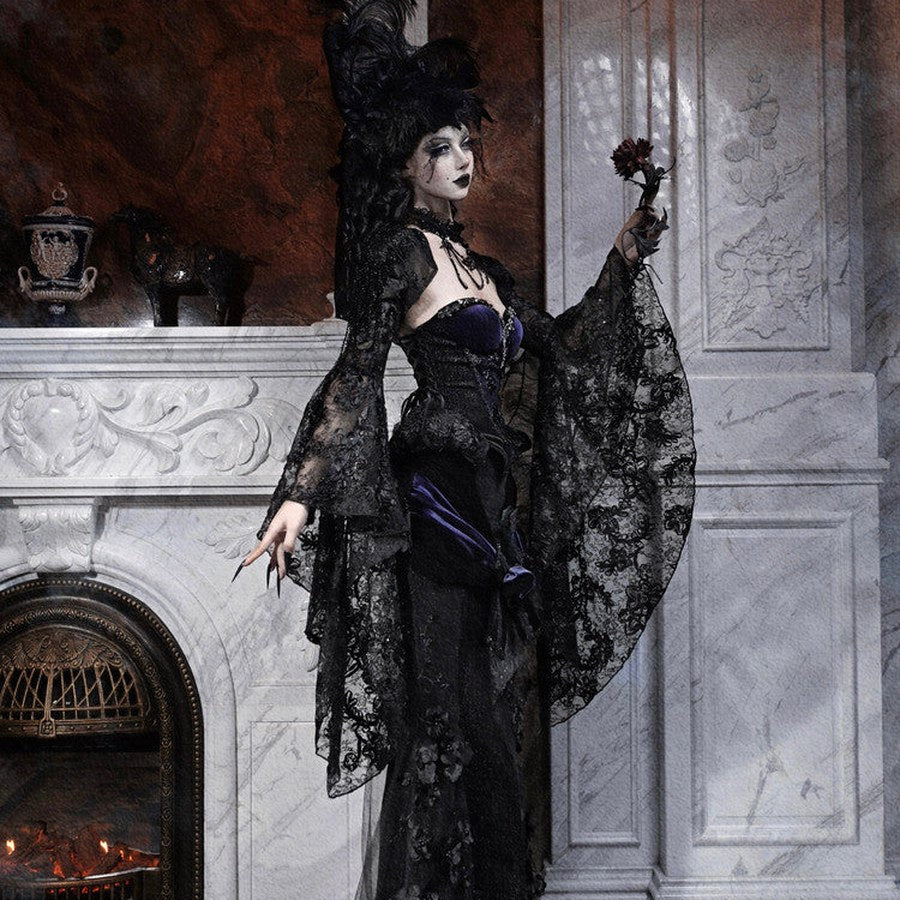 Model in a gothic black lace robe with purple corset, feathered hat, holding a small dark rose by a fireplace.