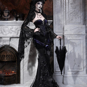 Model in a gothic black lace mermaid skirt with purple corset, holding a closed black umbrella by a fireplace.