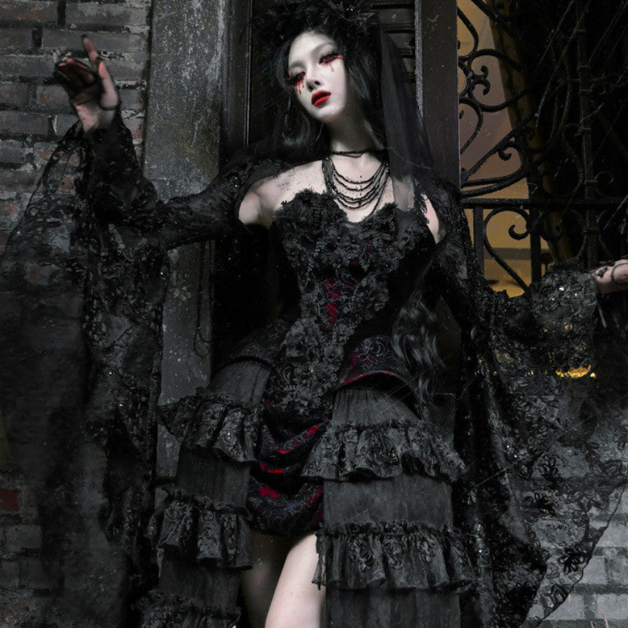 Gothic black velvet corset bustier top with sheer lace cape and tiered ruffled skirt, outdoors against stone wall backdrop.