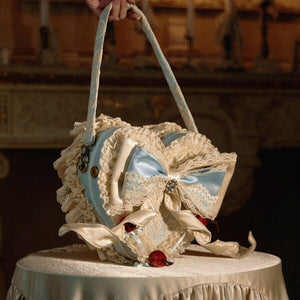 Blue heart handbag angled on a cream table showing lace trim, side depth, and shoulder handle