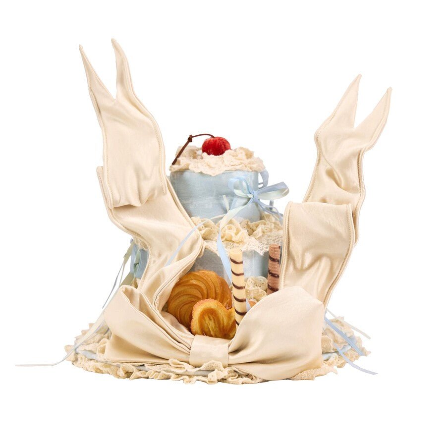 Flat lay of the Cookie Cake Ribbon Hat item with satin wing details, lace trim, and dessert accents
