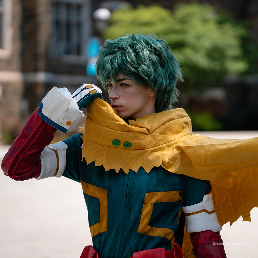 Deku cosplay costume upper-body close-up frames a yellow hooded cape over teal suit panels and a white wrist guard.