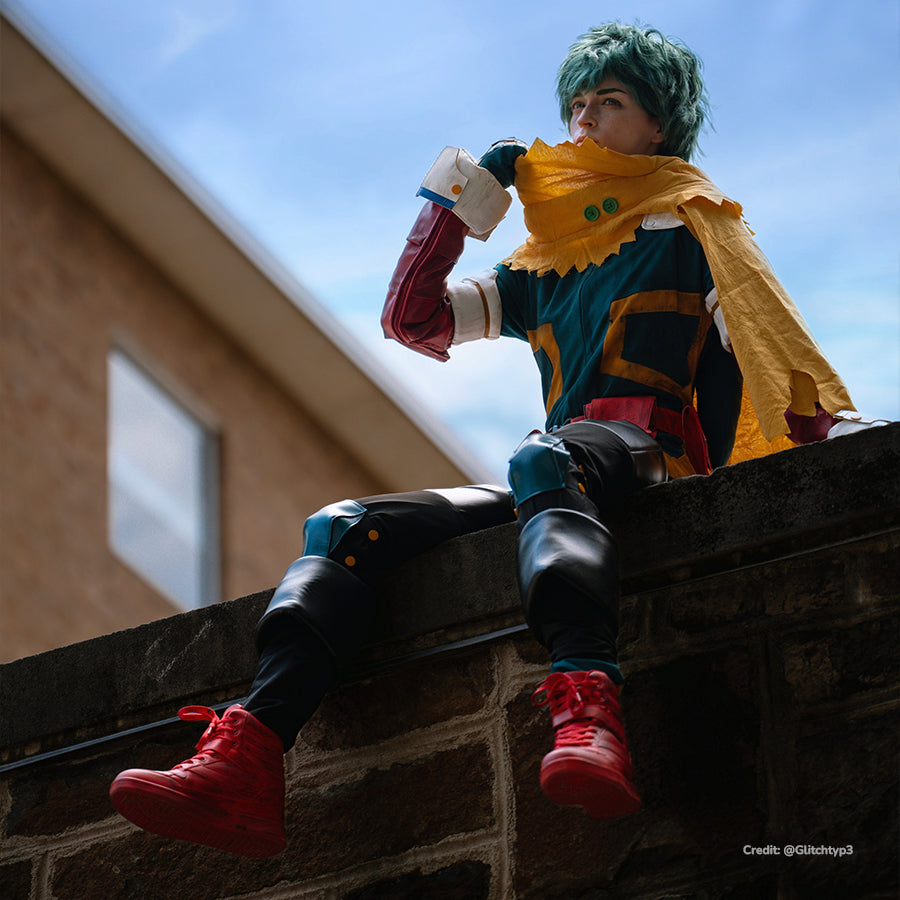 Deku cosplay costume seated view sets a yellow hooded cape above teal suit panels and black knee guards.