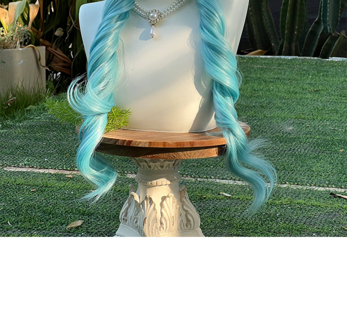 Close-up view of peacock teal blue lolita wig showing long braided pigtails on a mannequin outdoors, displayed on grass with potted plants and greenery in the background